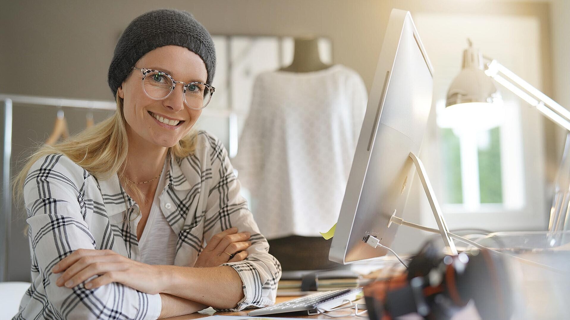 Eine junge Frau sitzt an einem Tisch mit einem Monitor und lächelt in die Kamera.